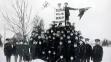 A crowd of soldiers are gathered around a pedestal, the top of which is visible under a vertical banner. The banner bears the phrase "Berlin will be Berlin No Longer". Two soldiers standing below and to the left of the bottom of the banner hold medallions showing the likenesses of Bismark and Von Moltke.