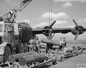 Black and white photograph of an aircraft on the ground with a group of men using a truck-mounted crane to lift a large tube-shaped bomb