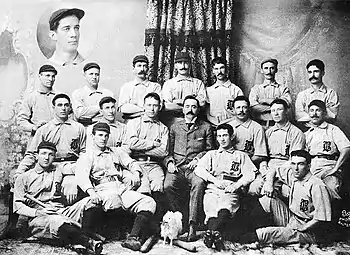 Three rows of men in white baseball uniforms and dark caps; the rear row is standing, the middle row is seated (with a man in a tweed suit in the middle), and the front row is seated on the floor. The baseball uniforms have a dark Old English-style "B" over the left breast.