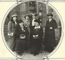 Six women wearing long winter coats and hats standing in front of a building