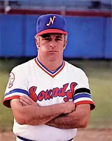 A man in a white baseball jersey with "Sounds" written on the front in red and blue and a blue cap with a white "N" on the front stands with arms crossed and a stony-faced expression.