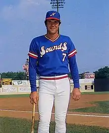 A baseball player in a blue baseball jersey with "Sounds" written on the front in red and white stands leaning on a baseball bat.