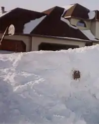 A large pile of snow piled up in front of a home, nearly obscuring it. A mailbox is peeking through the pile of snow and a basketball hoop can be seen behind the snow in front of the home. Snow is piled up in small areas on top of the home's roof.