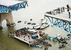 The picture is taken from a vantage point above the bridge disaster. The middle section of the bridge is missing, and spectators with umbrellas can be seen observing the collapsed section of the bridge which is floating in the water. Multiple small boats and kayaks can be seen crowding around the collapsed section of the bridge. There is a large crowd of people seen standing on the collapsed section of the bridge, along with a severely damaged bus, van, and car.