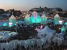 hundreds of spectators in front of the ice castle which is lit blue-green from inside