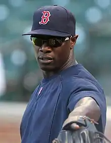 A man, wearing a blue baseball cap with the red letter "B" logo of the Boston Red Sox, points forward with the baseball glove on his left hand