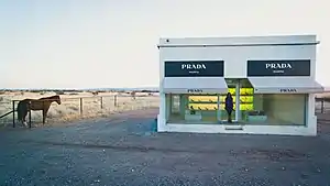 A small one story, one room adobe building with two large plate glass windows. Purses and shoes are sparsely and methodically placed inside.