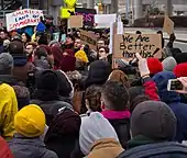 Crowd of protesters at John F. Kennedy International Airport. Visible signs read "America, land of immigrants", "Let them in", "Too mad to stay home", "We are better than this!"