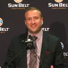 A color photograph of Jake Spavital in a suit and tie at a podium.