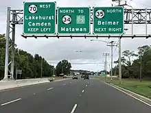 A six-lane divided highway approaching an intersection with a set of three green signs over the roadway. The left sign reads Route 70 west Lakehurst Camden keep left, the middle sign reads north Route 34 to Garden State Parkway Matawan, and the right sign reads Route 35 north Belmar next right.