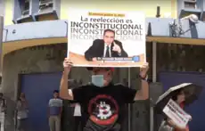 A street protesters holding a sign with a quote attributed to Armando Bukele Kattán reading "Re-Election is Unconstitutional" in Spanish.