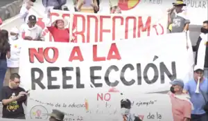 Street protesters holding a white banner with red text reading "No to Re-Election" in Spanish.