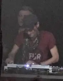 A DJ with a red shirt and black cap performs live on a stage, with former Underworld member Darren Emerson standing behind him