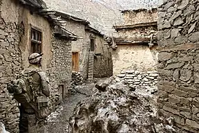 A corpsman in desert fatigues stands in the middle of a group of very old houses.