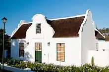 This house was probably designed by the well-known Thibault and built in 1815 for the widow of the Reverend H. W. Ballot. The strange geometric gable is exceptional. Elizabeth Wilhelmina Cruywagen, widow of Ds H. W. Ballot, after her husband's death in 1814, applied for a piece of land to build a house – she had to vacate the parsonage – all land E of Church St had already been sold, subdivided and built upon. Therefore, land was allotted on the W side of the street, opposite the pastorie, and surveyed by the younger Schutte – the boundary runs through a corner of the house. The actual grant is dated 1818. The house is U-shaped, but differs in many ways from others of its time. It has its long back-wings close together, with space only for a 2,8 m wide passage, which was roofed over from the beginning, and is the continuation of the entrance passage – the house never had a wide voorhuis, but may have had screen-like double doors with glazed upper parts. The narrow front elevation consisted of a vertically divided front door with only one large sash window on either side (this elevation was later completely changed, but could be accurately reconstructed, also because the original sliding sashes were discovered by Dr Mary Cook, present owner of the house, on a farm nearby). The great height of the ceilings in the house is also unusual. Though the side gables are of the traditional holbol type and those at the back are straight, the front gable, which in spite of the many later alterations was never removed, is unlike anything else in Church St. It has a central projecting section, crowned with a wide, segmental pediment like that of Groot Constantia, but with concave-convex lower outlines. The string-course is sharply stepped up at the ends. Part of the gable face had grooves to suggest ashlared stonework. (The date 1815 used to be faintly legible under the red paint with which the whole facade was at one time covered.) All these features suggest that a designer more sophisticated than those responsible for the other Church St houses (except, of course, no 36) was involved in building Ballotina. Who was he? Louis-Michel Thibault, the architect of the Drostdy, must have been a frequent visitor to Tulbagh during the years 1805-1807. Later, in 1812, he did work there as a land-surveyor. His wife was both a first cousin and sister-in-law of Mrs Ballot, and he could have stayed nowhere but at the Parsonage whenever he was in Tulbagh. After Ballot's death he may have provided her with a stock plan, to be carried out by local builders (he died in the year the house was built), or else he may already have given it to Ballot himself, to be used for a house to retire in. (For the pediment, cp that of the Drostdy and for the plan, cp Saasveld.) From 1870 till 1905, Ballotina was used by the State School founded by the Divisional Council. The house was restored in 1972. Type of site: House Previous use: Residential. Current use: Residential.