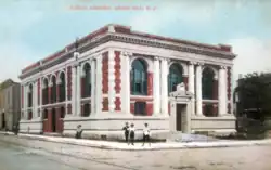 An old postcard of a library on a street corner, where a man is walking by with a top hat.