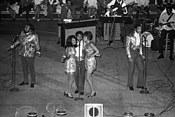 Three dark-skinned men and two dark-skinned women singing on a stage.  Various musicians are visible behind them.