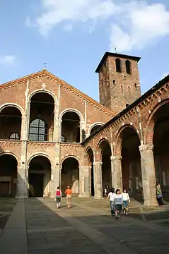 The façade and forecourt of a redbrick church are composed of simple arcades. A brick tower rises up to one side.