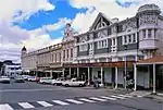 Splendid Neo-Baroque facade. Steeply pitched pediment plus corner turret. Heavy modulated cornice and frieze. Coupled columns and ranges of windows. Original shop front missing. Destroyed in fire of 1906. Rebuilt in 1907 to design of H. Walker – Port Elizabeth. See c5 in pilot study. Type of site: Commercial. Current use: Commercial – Shops. Late Victorian/Edwardian double storey shop, forming part of important grouping.