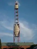 A red lattice tower with decorative yellow panels in a zig-zag diamond shape and more panels up top. A light panel sits just below the top of the mast.