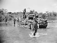 A tank moves across a shallow creek with armed soldiers advancing beside it