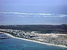 View across a sandy strip of land, with ocean on both sides. The land nearest the observer is mostly sandy, and has many buildings on it, and some jetties. The land furthest from the observer is thickly vegetated. The ocean in the foreground is blue-green, with dark patches indicating areas of coral reef. The ocean in the background is light blue. There is a line of breakers in the distance.