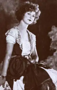 A white woman with curly bobbed hair, seated, wearing a dress with short sleeves and lace trim