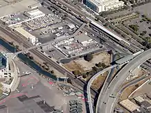 Aerial view of a station complex with three platforms in an industrial area. A footbridge connects the platforms to a stadium.