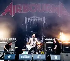 Three members of the band are in view. The band's name 'Airbourne' is displayed on a back-board, above the drummer, who is obscured behind the lead singer.