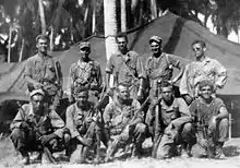 Black-and-white image of ten men in two rows, the top row standing and the bottom row crouching, are all facing the camera. They are wearing military attire and are holding rifles.