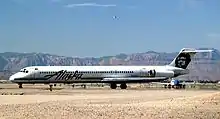 Right side view of an airplane taxiing on the ground towards left side of image. Another plane is behind it, and in the background are mountains and blue sky with a few clouds.