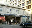 A light grey building with "M.R. ALDGATE STATION M.R." written in stone on the front face and a black car driving in the foreground