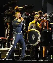 A man dressed in Mexican charro suit, holding a microphone in his left hand and a hat on the right.