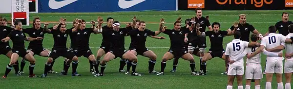 The New Zealand team lined up, with their arms raised to their side and palms facing down, mouths open in full voice, and eyes looking directly at their opponents opposite. The New Zealanders are wearing black shorts and socks, while squatting with knees bent and backs straight.