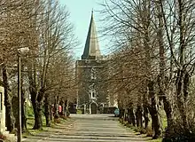 A tree lined road leads to a grey stone tower topped by a spire