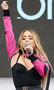 Brooke, dressed in a pink and black long-sleeve outfit, sings into the microphone.