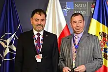 Two older European men in suits and ties stand in front of flags.