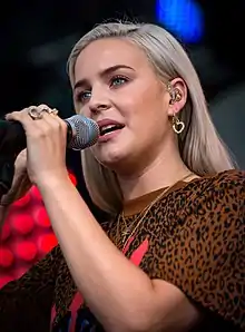 blonde white female in front of microphone, grinning with her one hand up to her ear