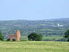 Patchwork of fields and trees with buildings showing in the distance. In the foreground is grass with a ruined building.