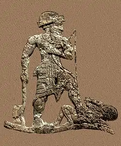 King Anubanini. He is equipped with an axe, a bow and an arrow. He is bare-chested, wears a short skirt, a roll-brimmed hat and sandals.