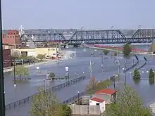 A street with a major amount of water on it due to flooding. A bridge is on the top of the image, and a row of buildings to the left. Sandbags are in front of the buildings