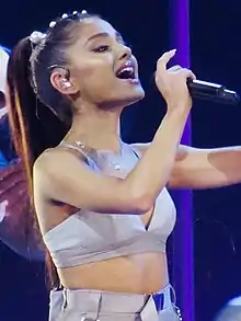 A woman with brown hair singing on stage wearing a lilac crop top and skirt