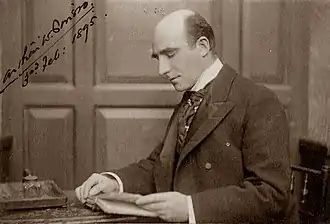 photograph of middle-aged white man, bald, clean-shaven, sitting at writing-desk