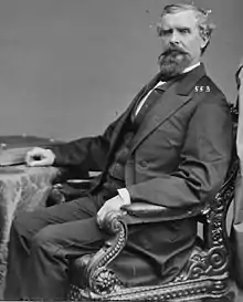 A man with graying hair, a beard, and a mustache, sitting, wearing a black jacket and tie and white shirt