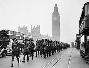 Black and white photograph of a large number of men wearing military uniforms marching down a street. A tower with a clock on it is visible in the background.