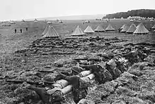 Black and white photo of eight men wearing military uniforms standing in a trench and aiming rifles. Tents are visible in the background.