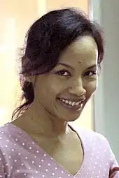 An Indonesian woman, looking forward and smiling. She is wearing a purple v-neck shirt