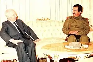 Saddam Hussein in uniform and man in suit, seated at opposite ends of a sofa and talking