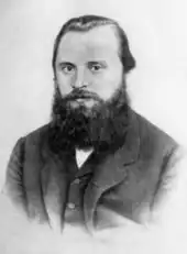 A man in his late 20s or early 30s with dark hair and a bushy beard, wearing a dark coat, dress shirt and tie