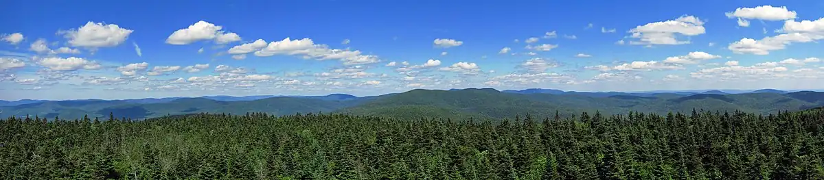 A forested expanse of mountains under a blue sky with fluffy white clouds